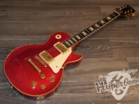 Red Les Paul–style electric guitar with gold hardware resting on a wooden floor.