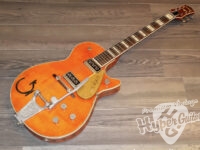 Orange electric guitar lying on a wooden floor with a Bigsby tailpiece and two humbucker pickups; Vintage-style sunburst finish and dark fretboard.