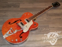 Orange Gretsch hollow-body electric guitar with white pickguard and Bigsby tailpiece resting on a wood floor.