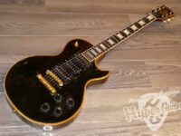Black electric guitar with gold hardware resting on a wooden floor, body facing the left side of the image on a light-brown plank floor.