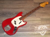 Red electric bass guitar with a white pickguard resting on a wood floor, angled diagonally across the image.