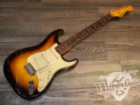 Sunburst Fender Stratocaster electric guitar with a white pickguard lying on a wood floor, showing its wear and chrome hardware.