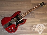 Red Gibson-style electric guitar with black pickguard on a wooden floor; chrome bridge and multiple knobs visible.
